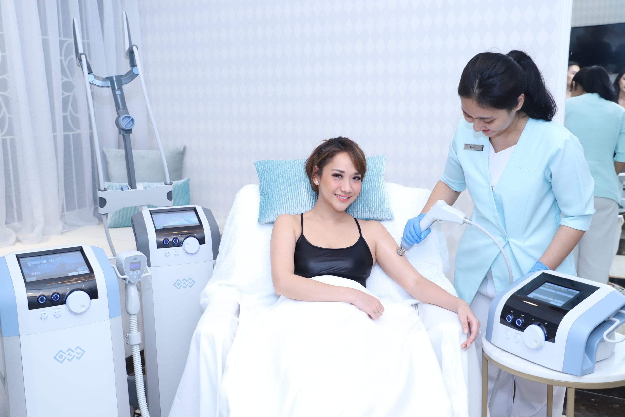 Bunga Citra Lestari wearing a black dress sitting on a treatment bed at Blink Beauty Clinic while getting a coolsulpting treatment from a therapist.