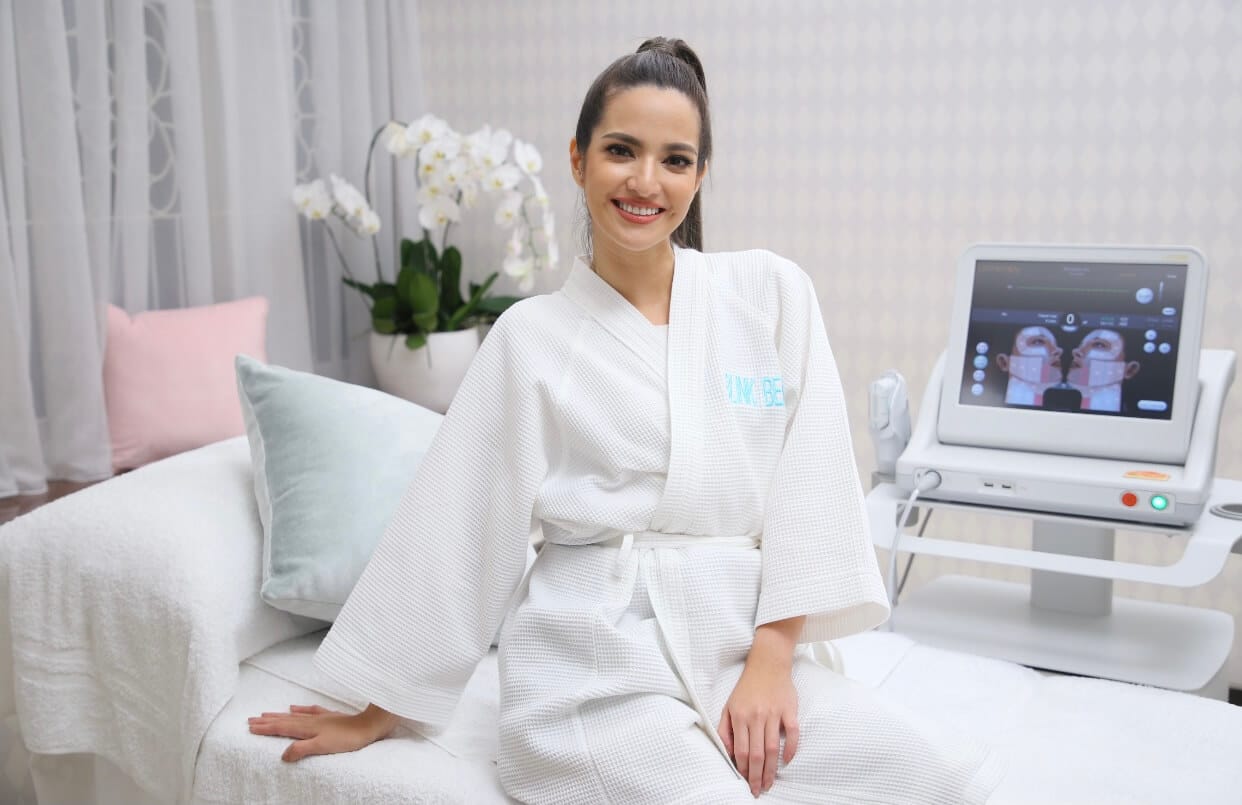 Nia Ramadhani in a white robe at Blink Beauty Clinic sitting on a treatment bed smiling at the camera before her treatment.