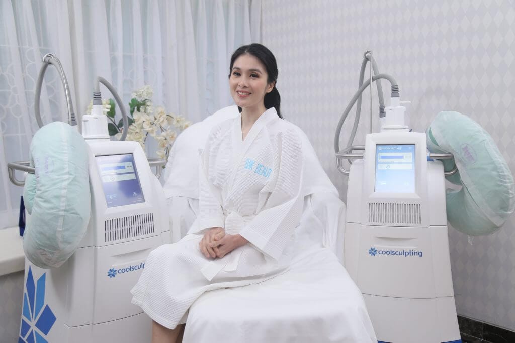 Sandra Dewi in a white robe sitting on a treatment bed at Blink Beauty Clinic smiling next to two coolsculpting treatment machines.