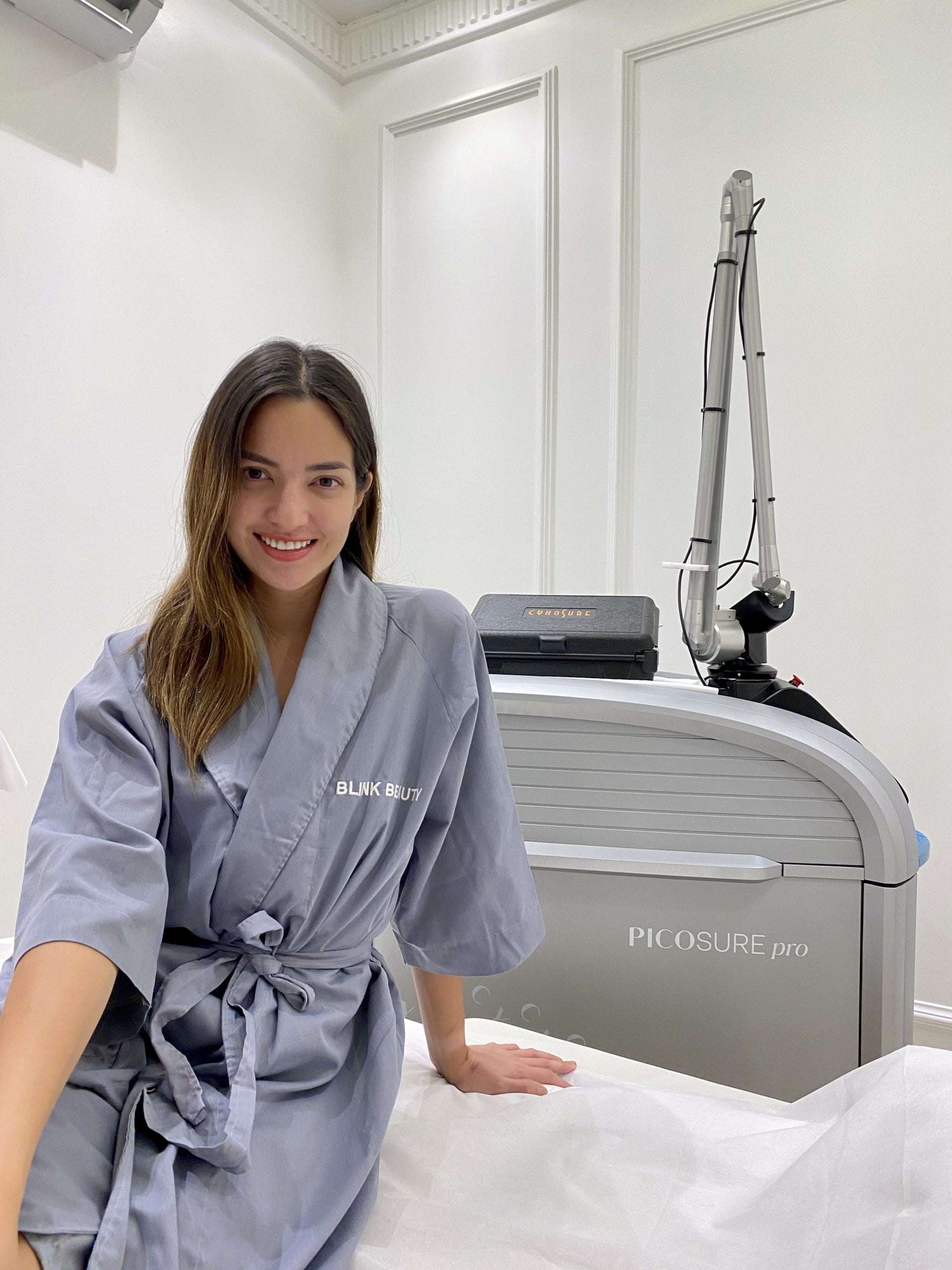 Nia Ramadhani in a gray robe at Blink Beauty Clinic sitting on a treatment bed smiling at the camera before her Picosure Pro treatment.