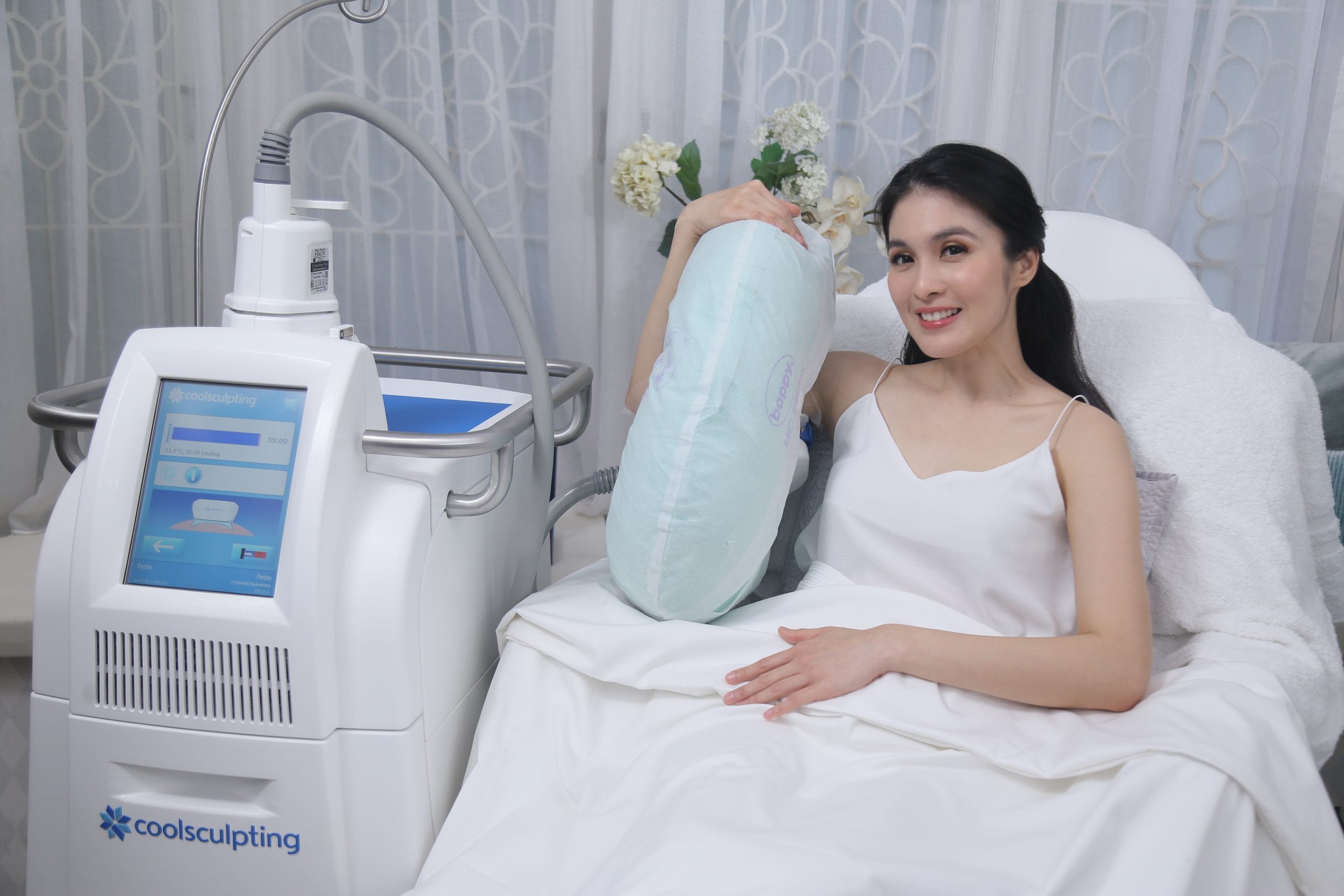 Sandra Dewi in a white robe sitting on a treatment bed at Blink Beauty Clinic smiling next to a coolsculpting treatment machine.