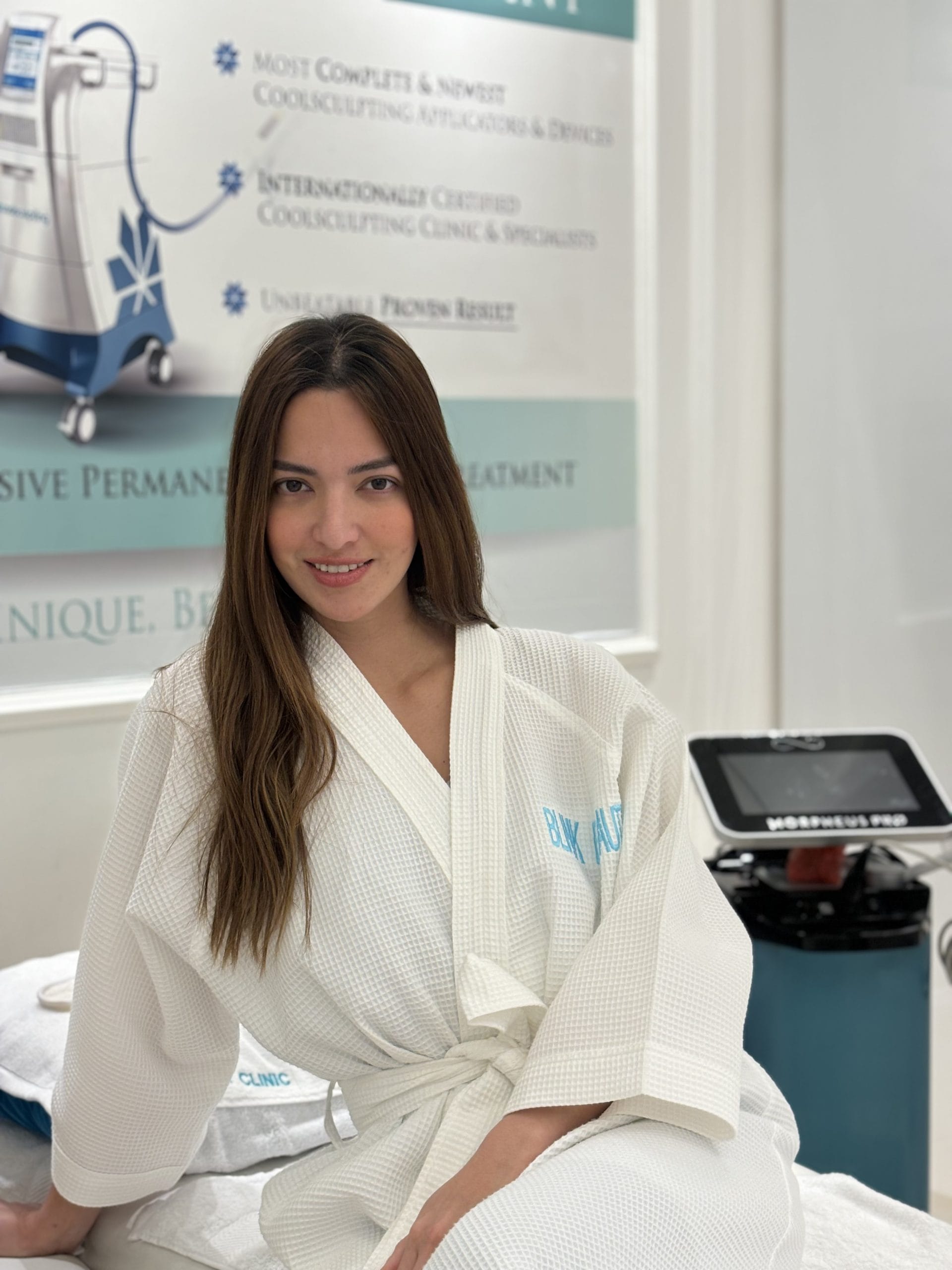 Nia Ramadhani in a robe at Blink Beauty Clinic sitting on a treatment bed smiling in front of a morpheus neo machine.