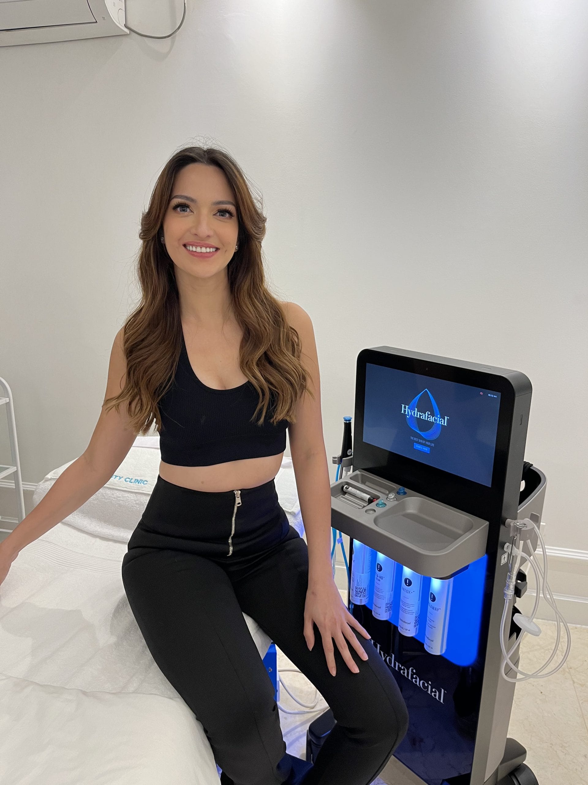 Nia Ramadhani at Blink Beauty Clinic sitting on a treatment bed smiling next to a Hydrafacial neo treatment machine.